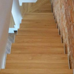 Schody System - Widok z góry na drewniane schody z białymi podstopnicami, przylegające do ceglanej ściany i białej ściany, w nowoczesnym wnętrzu.