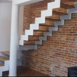 Nowoczesne schody z drewnianymi stopniami i białymi metalowymi wspornikami zamontowane przy ceglanej ścianie w Radomiu, z widoczną kolumną podtrzymującą i fragmentem pieca w dolnym rogu.