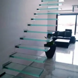 Nowoczesne schody z przezroczystymi stopniami szklanymi, wspornikowo mocowanymi do ściany, widoczne z boku w jasnym salonie z czarnymi meblami.