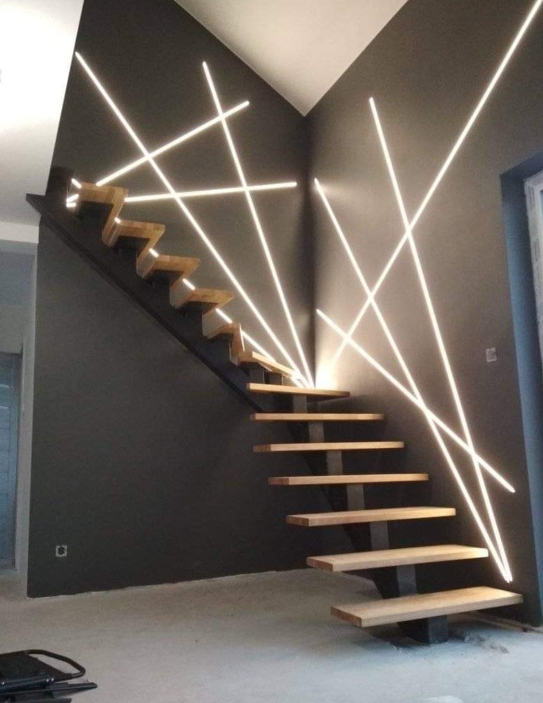 Nowoczesne schody z drewnianymi stopniami i metalową konstrukcją, podświetlone dekoracyjnymi liniami świetlnymi na szarej ścianie.