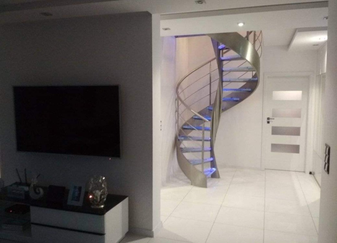 Nowoczesne, spiralne schody metalowe z podświetlanymi stopniami w jasnym wnętrzu, widok z dołu, obok ściana z telewizorem.