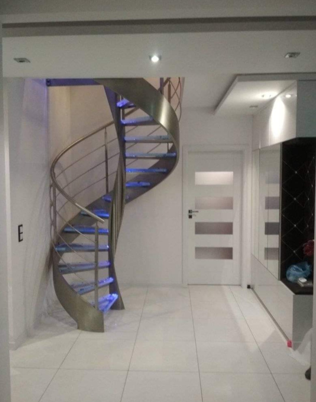 Wewnętrzne schody spiralne z metalu z podświetleniem LED, widok z dołu, minimalistyczny design, białe płytki podłogowe.