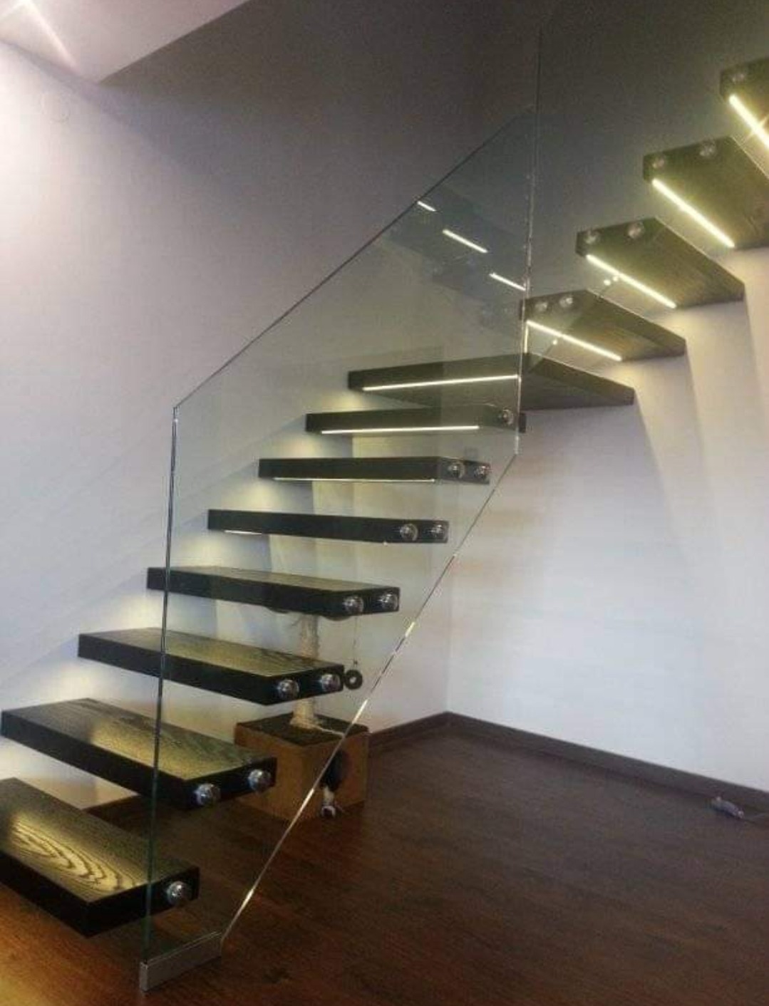 Nowoczesne schody z drewnianymi stopniami podświetlonymi LED i szklaną balustradą, wsporniki mocujące stopnie do ściany.