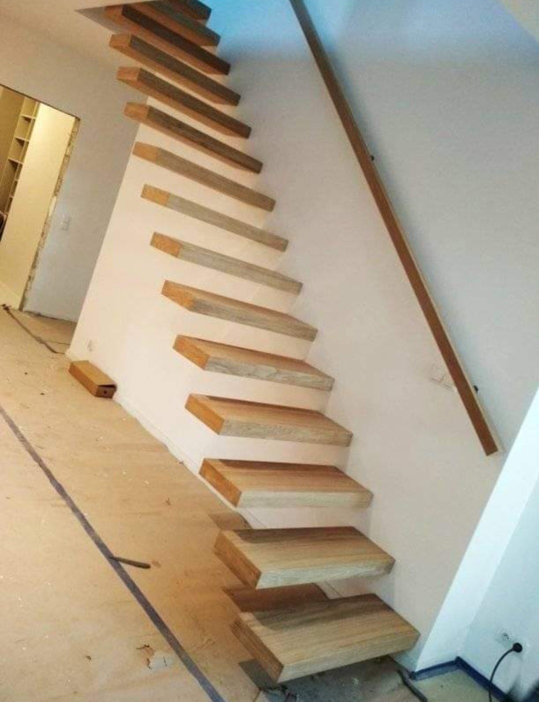 Nowoczesne, minimalistyczne schody drewniane wspornikowe z drewnianą poręczą zamontowane przy białej ścianie, widoczne ślady prac wykończeniowych na podłodze.