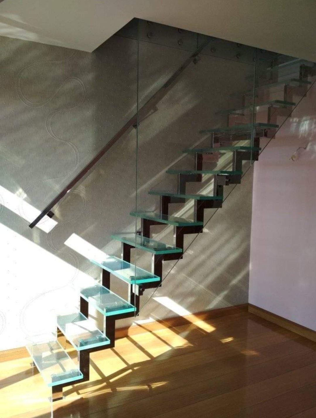 Szklane schody samonośne z drewnianą poręczą i szklaną balustradą, oświetlone promieniami słońca padającymi na parkiet i wzorzystą ścianę.