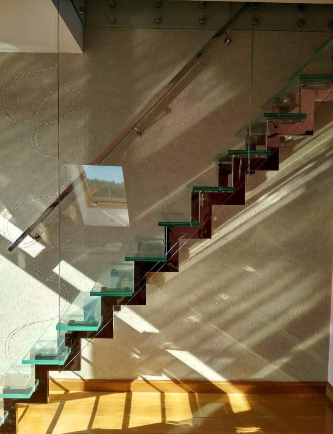 Nowoczesne schody z przezroczystymi stopniami szklanymi i metalową poręczą, oświetlone słonecznym światłem rzucającym cienie na ścianę z delikatnym wzorem.