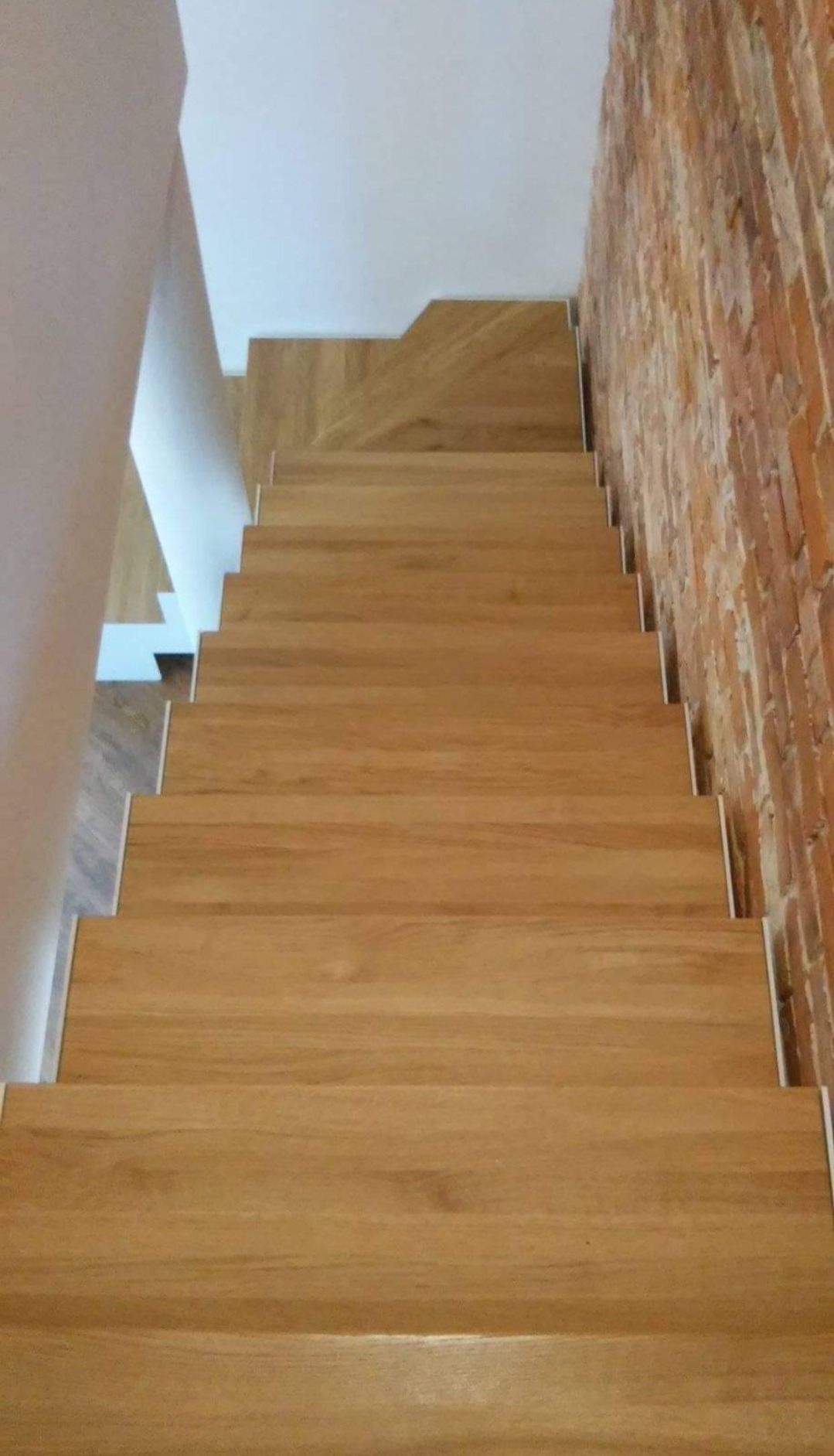 Widok z góry na drewniane schody z białymi podstopnicami, przylegające do ceglanej ściany i białej ściany, w nowoczesnym wnętrzu.