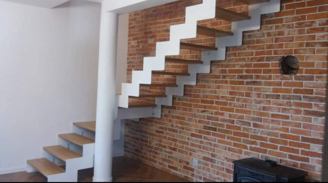 Nowoczesne schody z drewnianymi stopniami i białymi metalowymi wspornikami zamontowane przy ceglanej ścianie w Radomiu, z widoczną kolumną podtrzymującą i fragmentem pieca w dolnym rogu.