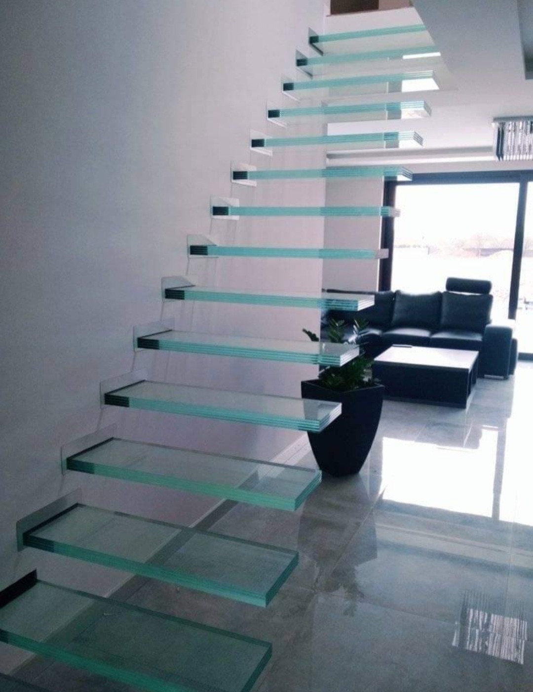 Nowoczesne schody z przezroczystymi stopniami szklanymi, wspornikowo mocowanymi do ściany, widoczne z boku w jasnym salonie z czarnymi meblami.