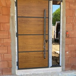 TurboMontażKraków Rafał Korepta - Montaż nowoczesnych drzwi wejściowych z drewnopodobnym panelem i długim uchwytem, widoczne niedokończone obramowanie z cegły i częściowo wypełniona pianką montażową ościeżnica, w tle zaparkowany bus.