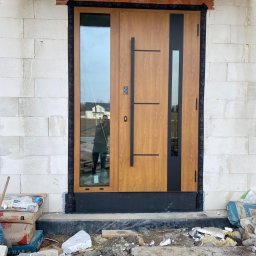 TurboMontażKraków Rafał Korepta - Nowoczesne, drewnopodobne drzwi wejściowe z czarnymi elementami, częściowo zamontowane w surowym murze z bloczków, widoczne materiały budowlane i odbicie fotografa w szybie.