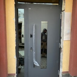 TurboMontażKraków Rafał Korepta - Montaż szarych drzwi wejściowych z długim uchwytem w folii ochronnej, widoczne uszkodzenia tynku wokół ościeżnicy i odbicie montażysty w szybie.