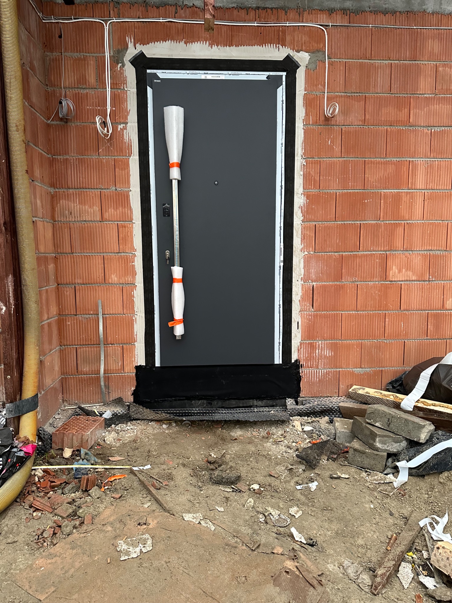 Montaż czarnych drzwi zewnętrznych w surowym murze z czerwonej cegły, z widocznymi przewodami elektrycznymi i zabezpieczoną klamką, otoczenie placu budowy.