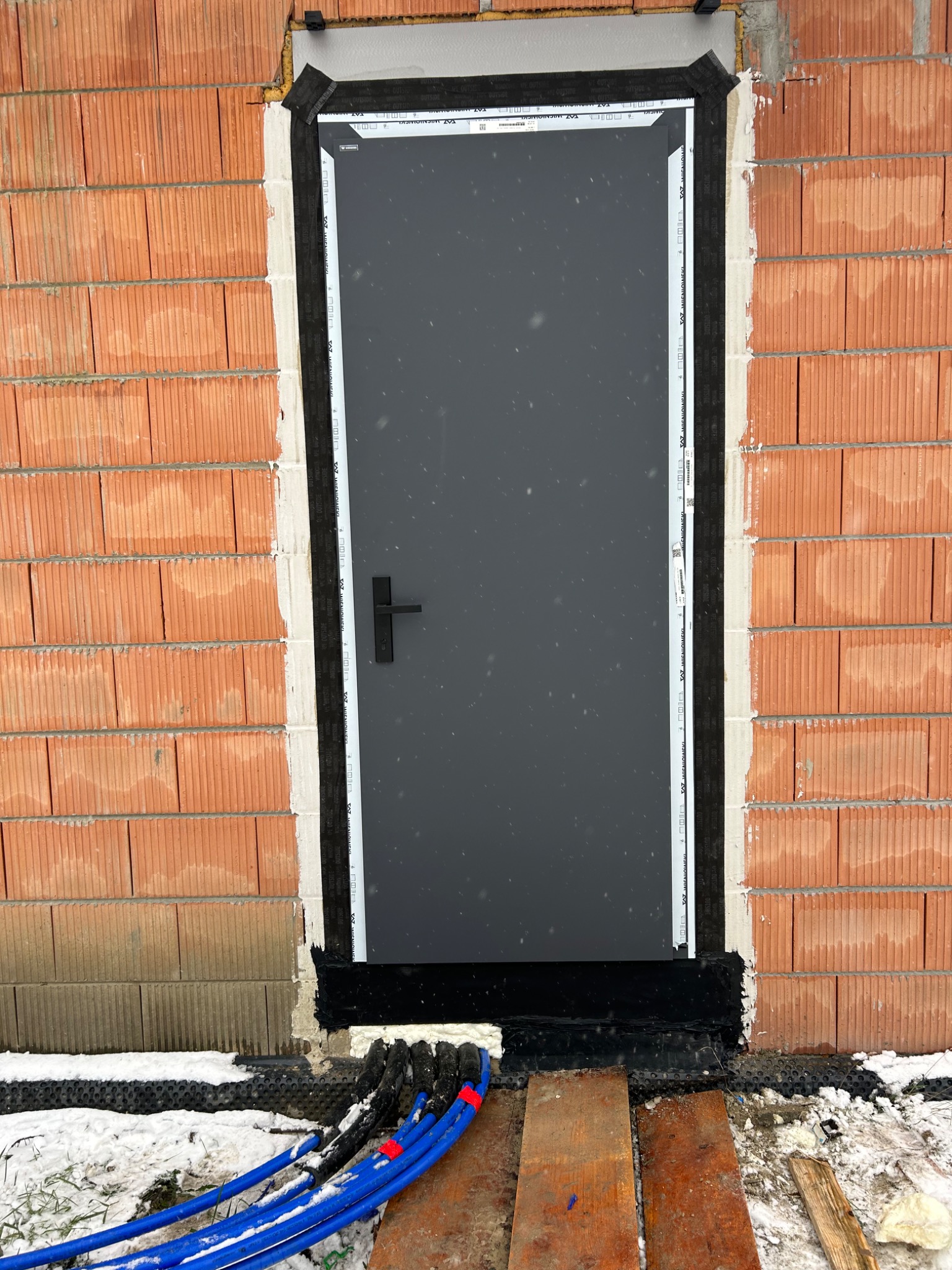 Nowo zamontowane, grafitowe drzwi zewnętrzne z czarną klamką, osadzone w surowej, ceglanej ścianie z widocznymi przewodami instalacyjnymi u dołu, częściowo pokryte śniegiem.