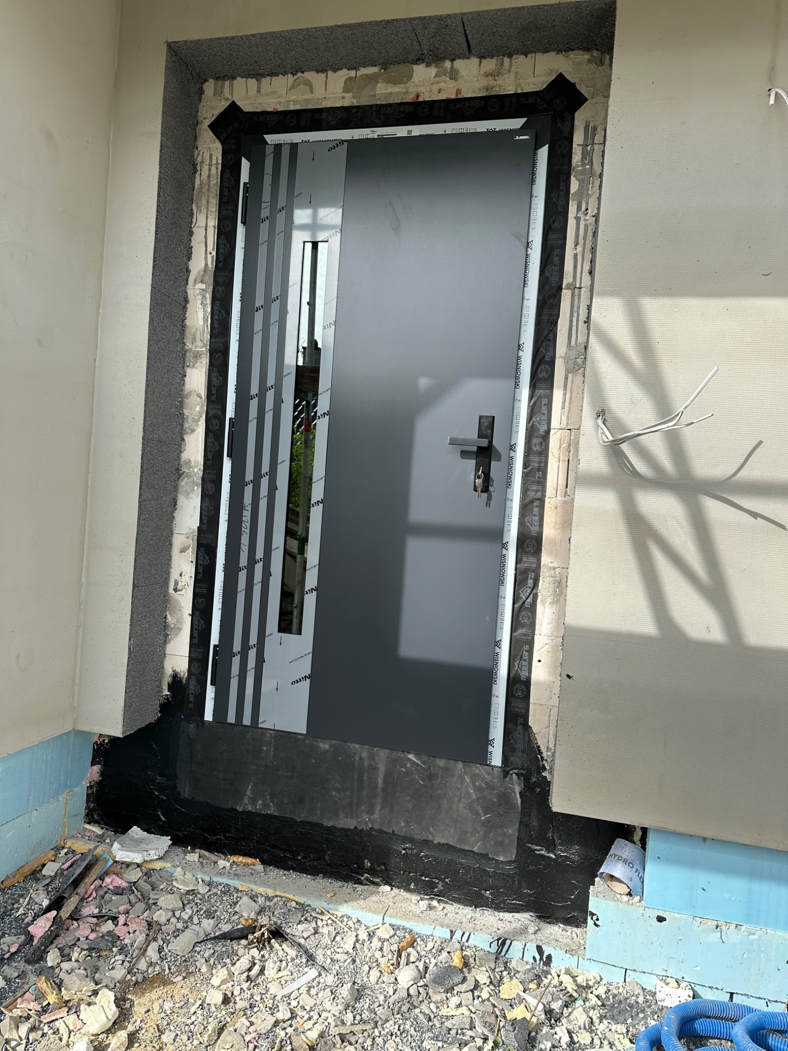 Szare drzwi wejściowe, zamontowane w surowym otworze, z widocznymi elementami montażowymi, uszczelnieniami i fragmentami gruzu na podłodze.