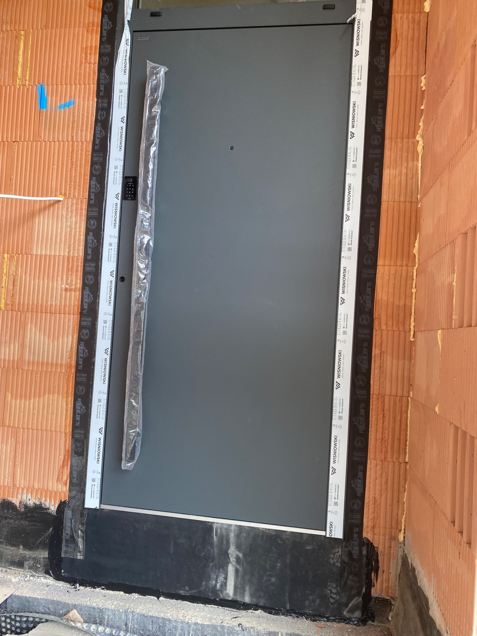 Szare drzwi zewnętrzne marki Wiśniowski w trakcie montażu, zabezpieczone folią, osadzone w murze z cegły ceramicznej, widoczne taśmy uszczelniające i pianka montażowa.