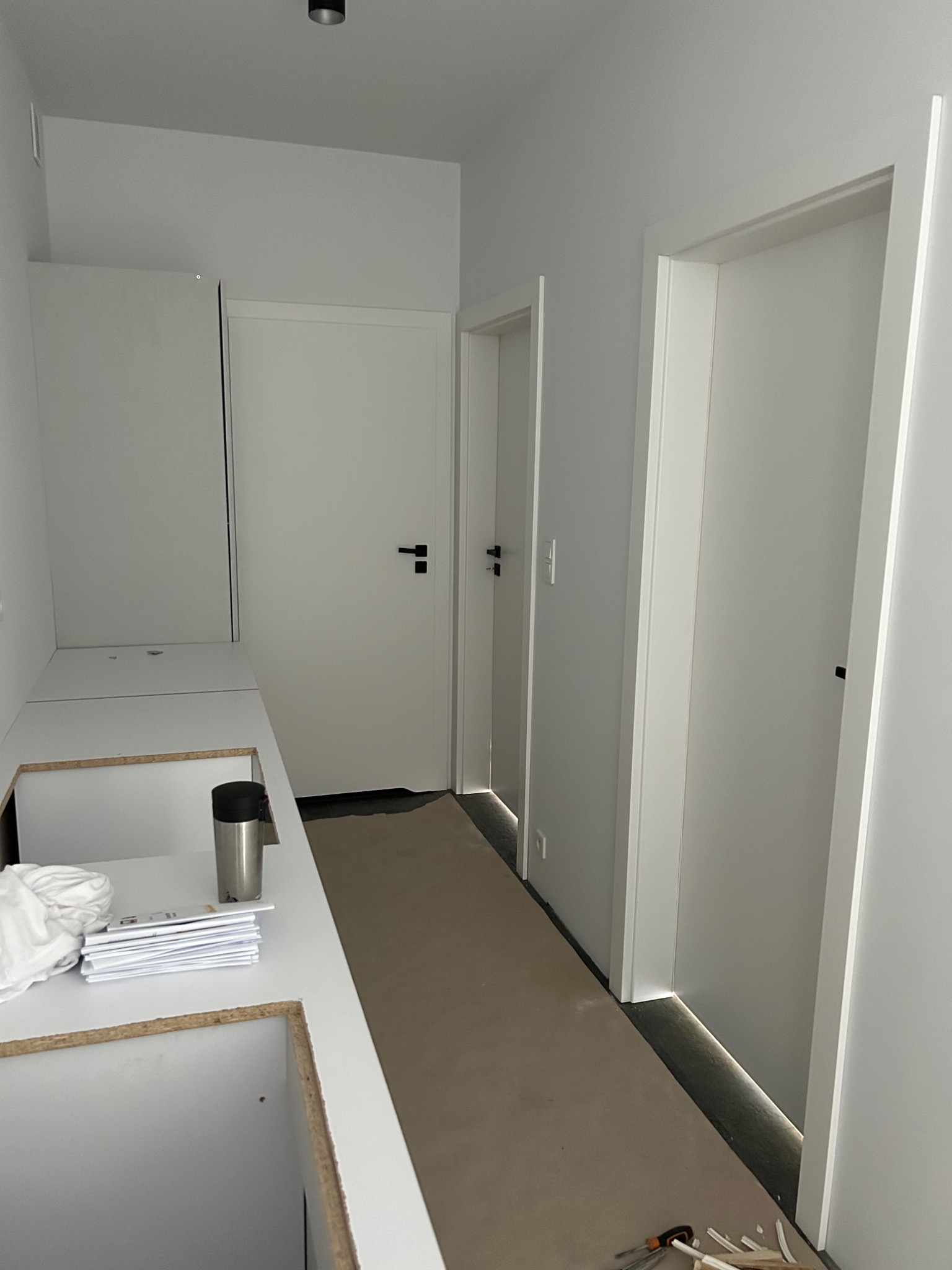 Wnętrze korytarza w trakcie wykańczania z trzema białymi drzwiami z czarnymi klamkami, widoczne narzędzia i materiały budowlane na podłodze, na blacie leżą dokumenty i termos.