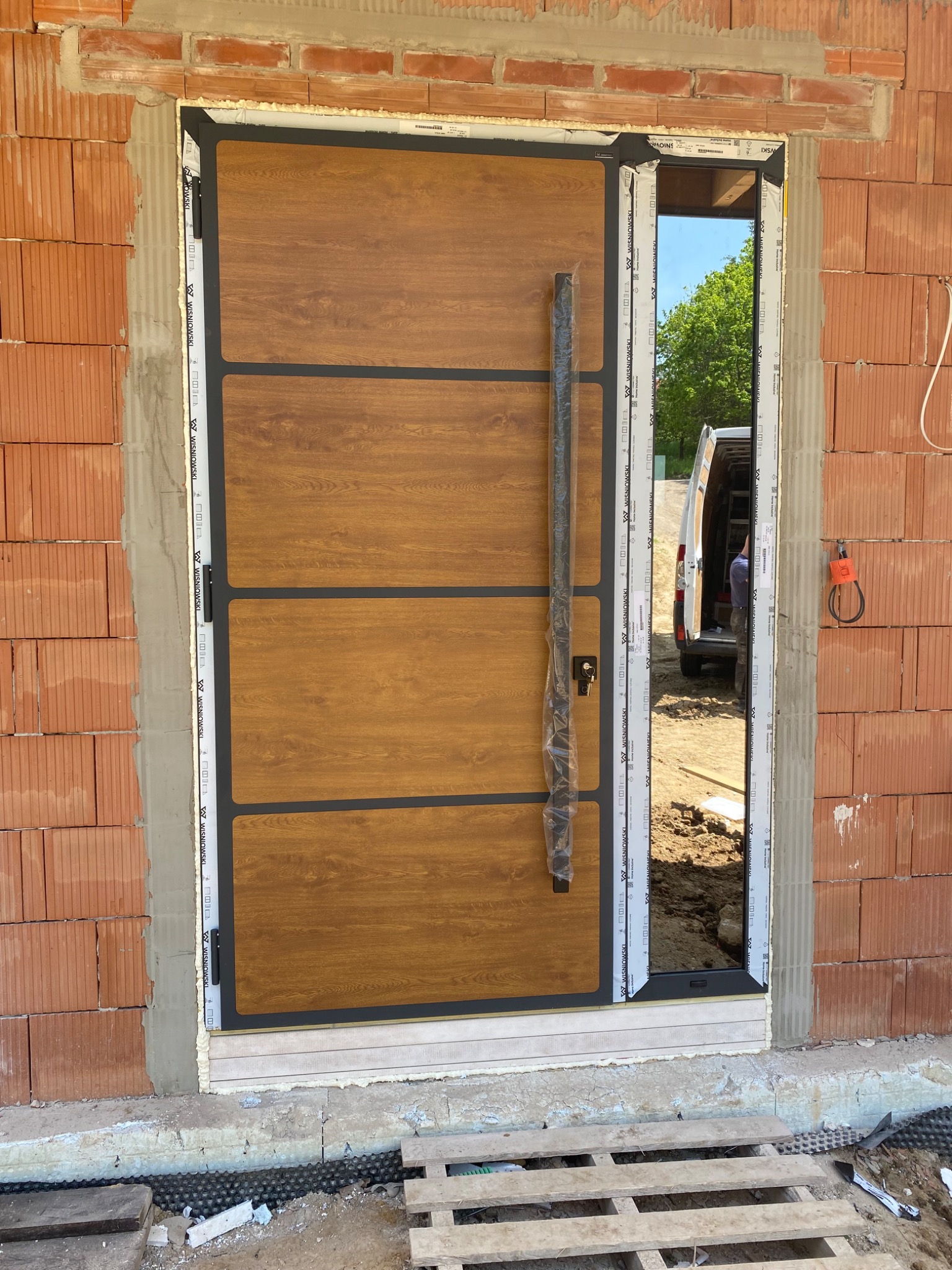 Montaż nowoczesnych drzwi wejściowych z drewnopodobnym panelem i długim uchwytem, widoczne niedokończone obramowanie z cegły i częściowo wypełniona pianką montażową ościeżnica, w tle zaparkowany bus.