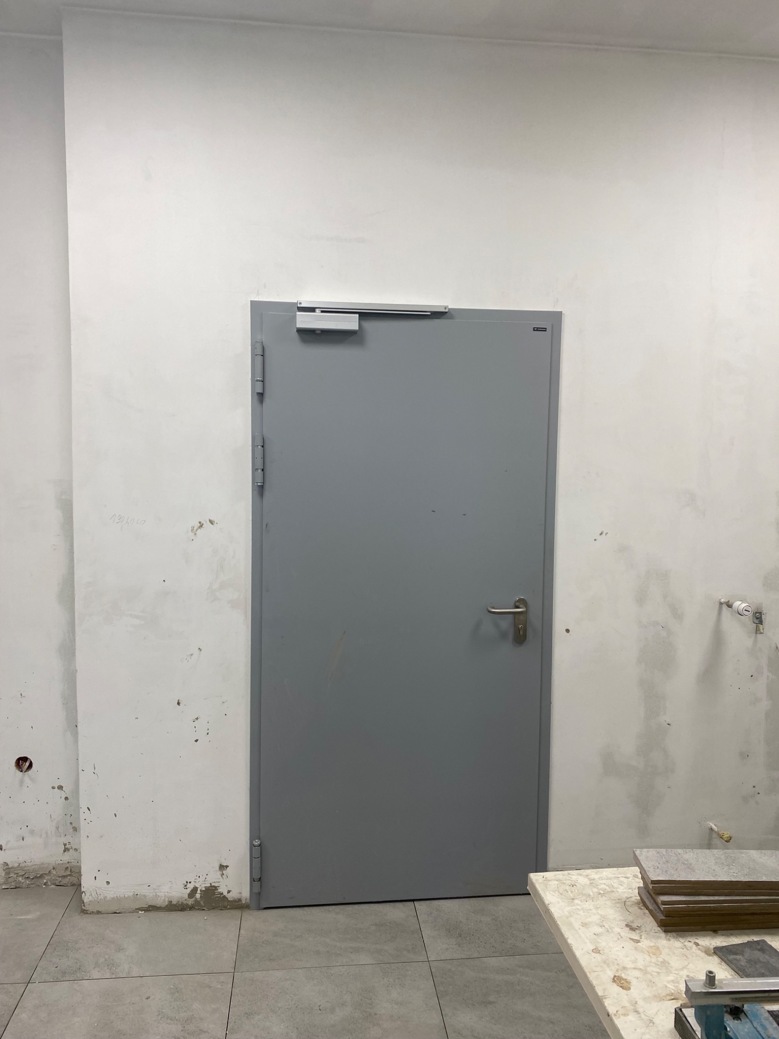 Szare, metalowe drzwi techniczne z samozamykaczem na tle surowej ściany w trakcie remontu, widoczne płytki ceramiczne na stole.
