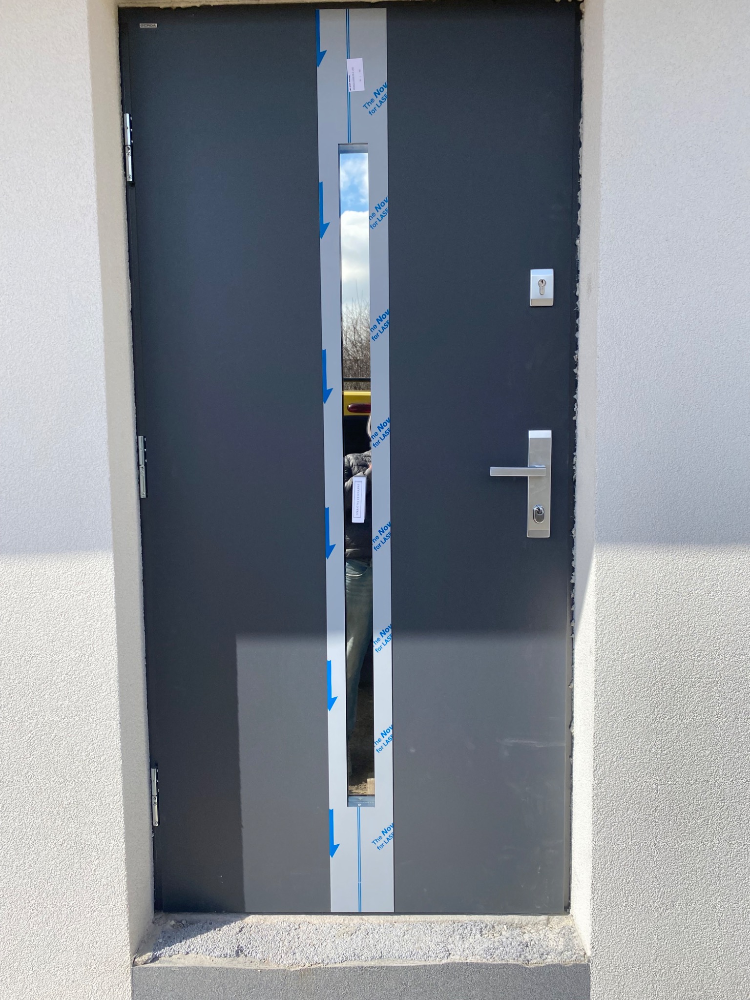 Ciemnoszare drzwi wejściowe z wąskim przeszkleniem zabezpieczonym folią ochronną, zamontowane w białej ścianie o chropowatej fakturze. Widoczny srebrny zamek i klamka.