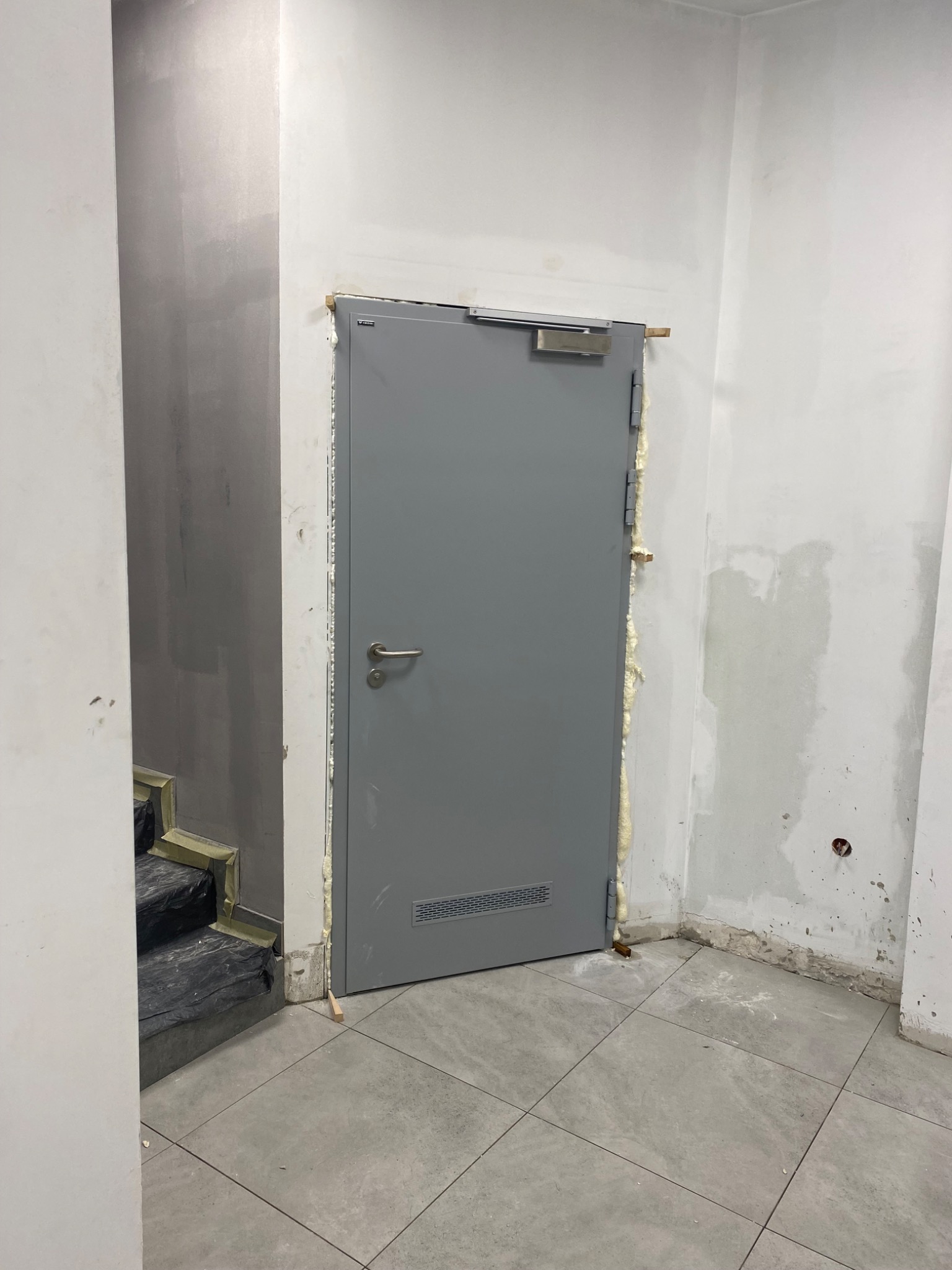 Szare, metalowe drzwi przeciwpożarowe, z widoczną pianką montażową wokół ościeżnicy, zamontowane w surowym wnętrzu klatki schodowej, z zabezpieczonymi schodami w tle.