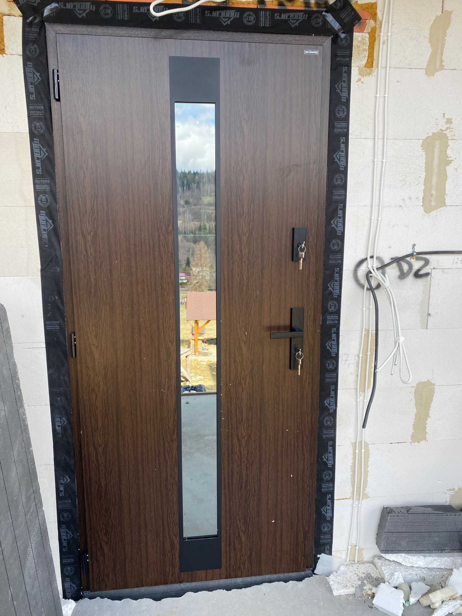 Nowo zamontowane, brązowe drzwi wejściowe z przeszkleniem pionowym, zabezpieczone taśmą montażową, widoczne klucze w zamku, surowy stan wykończenia ściany z instalacją elektryczną w tle.