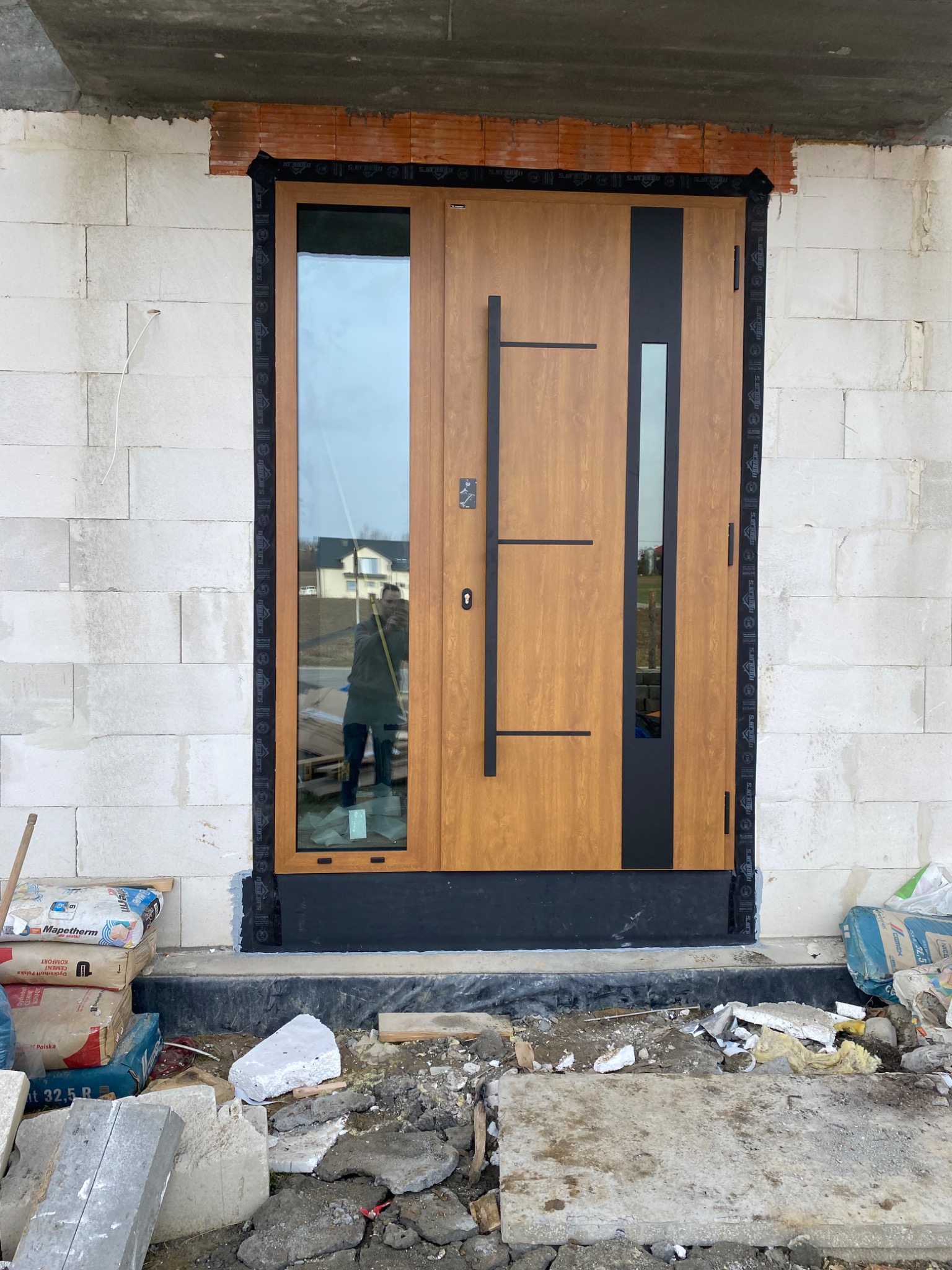 Nowoczesne, drewnopodobne drzwi wejściowe z czarnymi elementami, częściowo zamontowane w surowym murze z bloczków, widoczne materiały budowlane i odbicie fotografa w szybie.