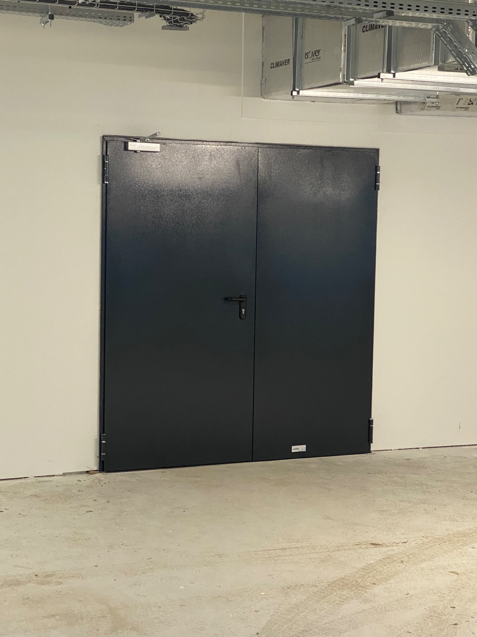 Zamknięte, dwuskrzydłowe, stalowe drzwi techniczne w jasnym pomieszczeniu, widoczny system wentylacji pod sufitem, zamek na środku.