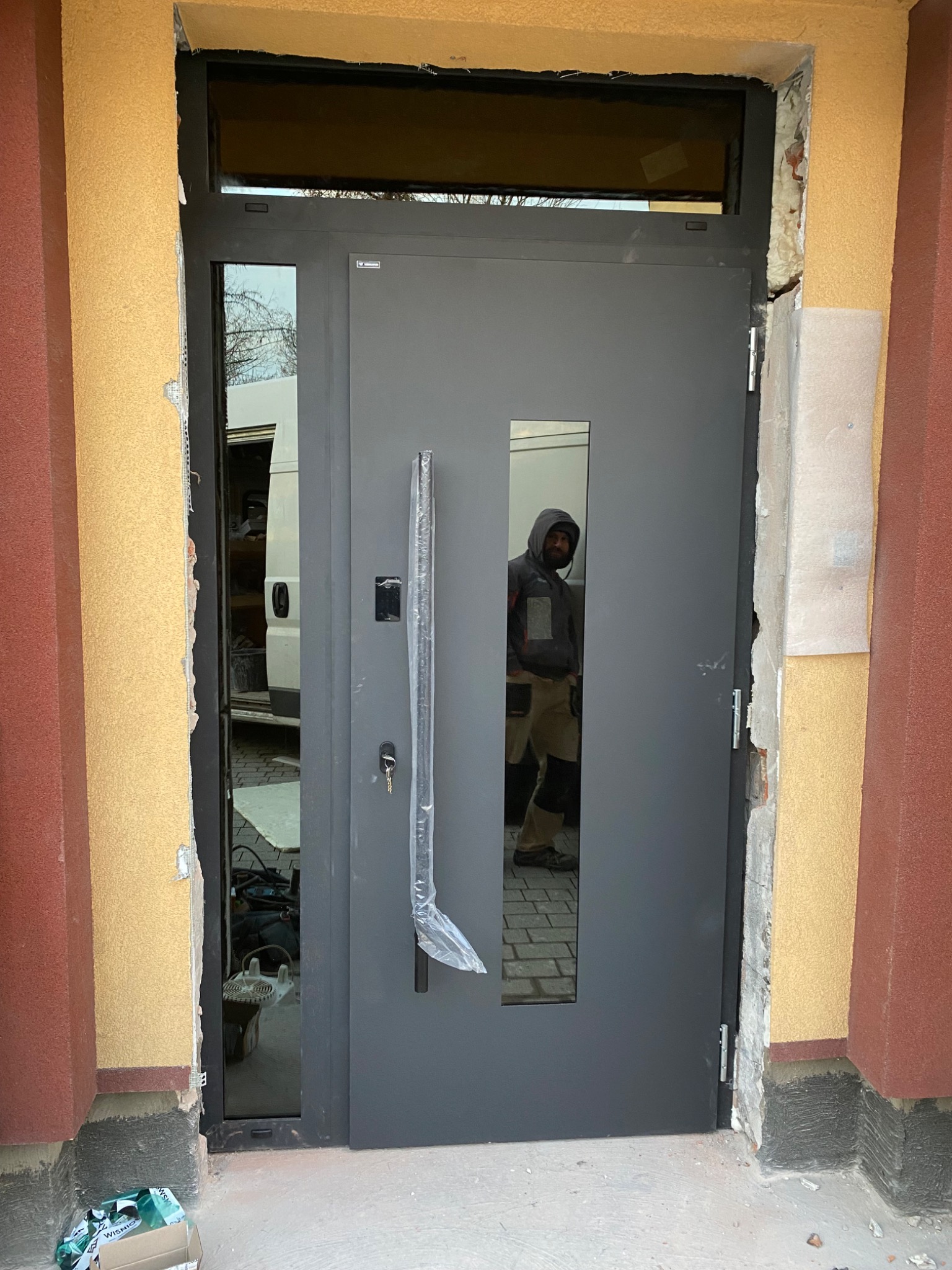 Montaż szarych drzwi wejściowych z długim uchwytem w folii ochronnej, widoczne uszkodzenia tynku wokół ościeżnicy i odbicie montażysty w szybie.