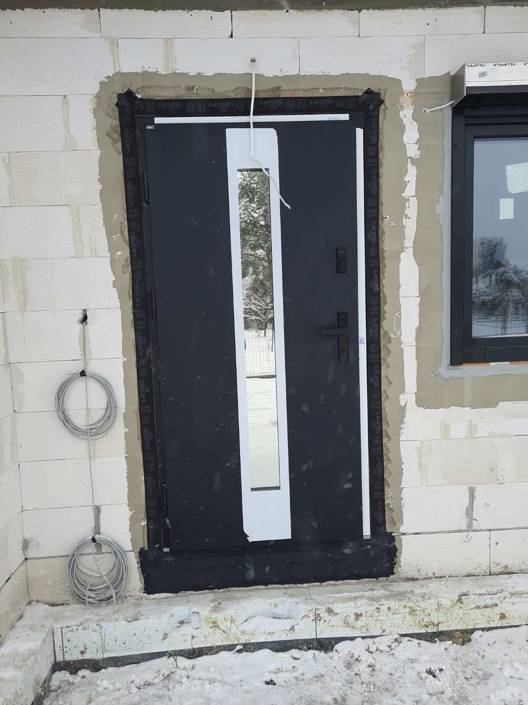 Czarne drzwi wejściowe z długim, pionowym, białym przeszkleniem, zamontowane w surowym murze z betonu komórkowego, z widocznymi śladami pianki montażowej i przewodami elektrycznymi na ścianie.