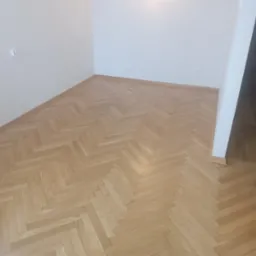 Jasny parkiet ułożony w jodełkę w rogu pokoju, widoczna ściana z gniazdkiem elektrycznym i fragment futryny drzwiowej.