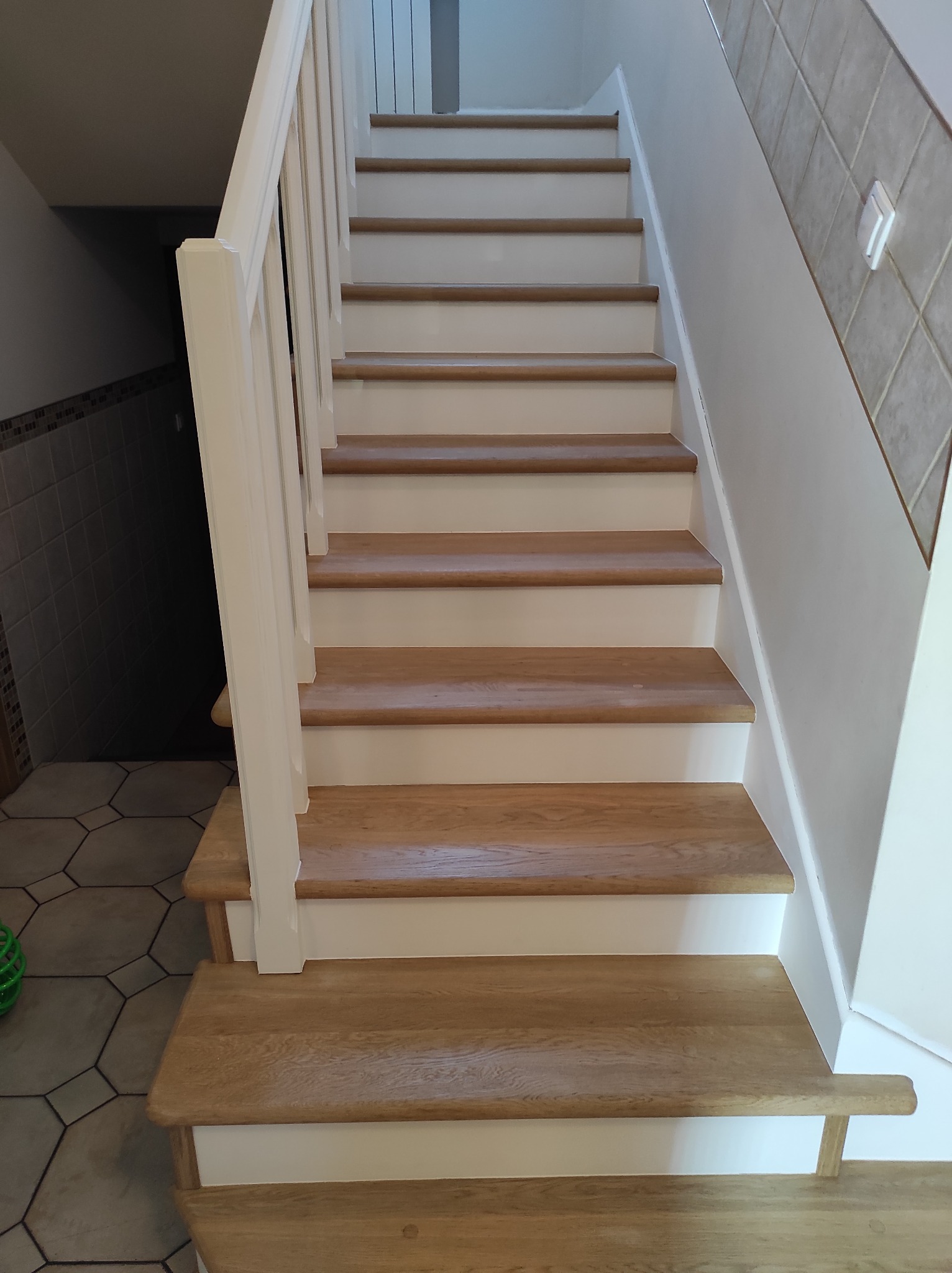 Odnowione schody drewniane z białymi podstopnicami i balustradą, widok z dołu, jasne drewno, płytki ceramiczne na ścianie i podłodze.