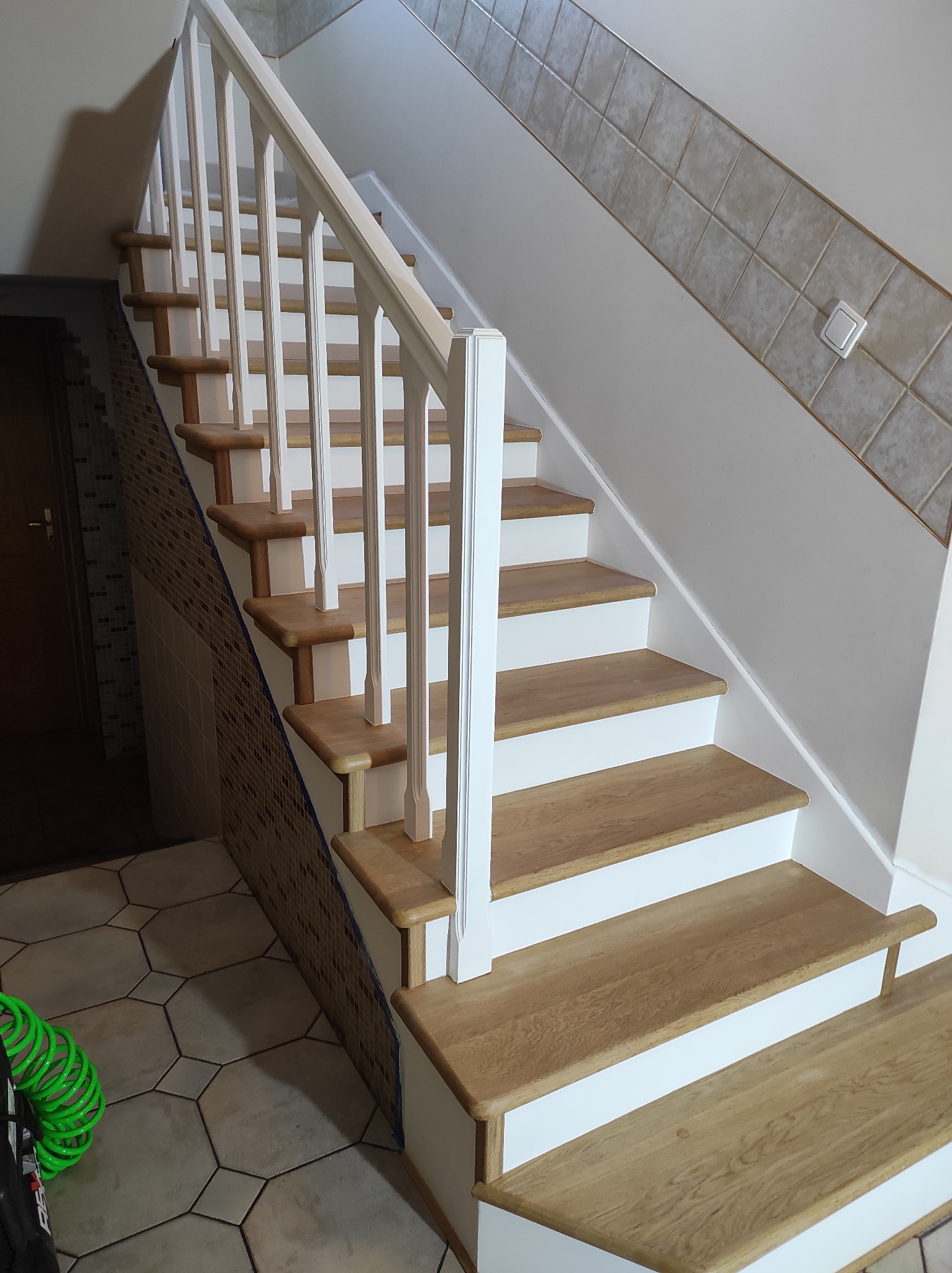 Odnowione drewniane schody z białymi podstopnicami i balustradą, widoczne płytki na ścianie i wąż pneumatyczny na podłodze.