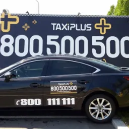 Czarny samochód TaxiPlus z numerami telefonów na karoserii, zaparkowany przed billboardem reklamującym aplikację mobilną.
