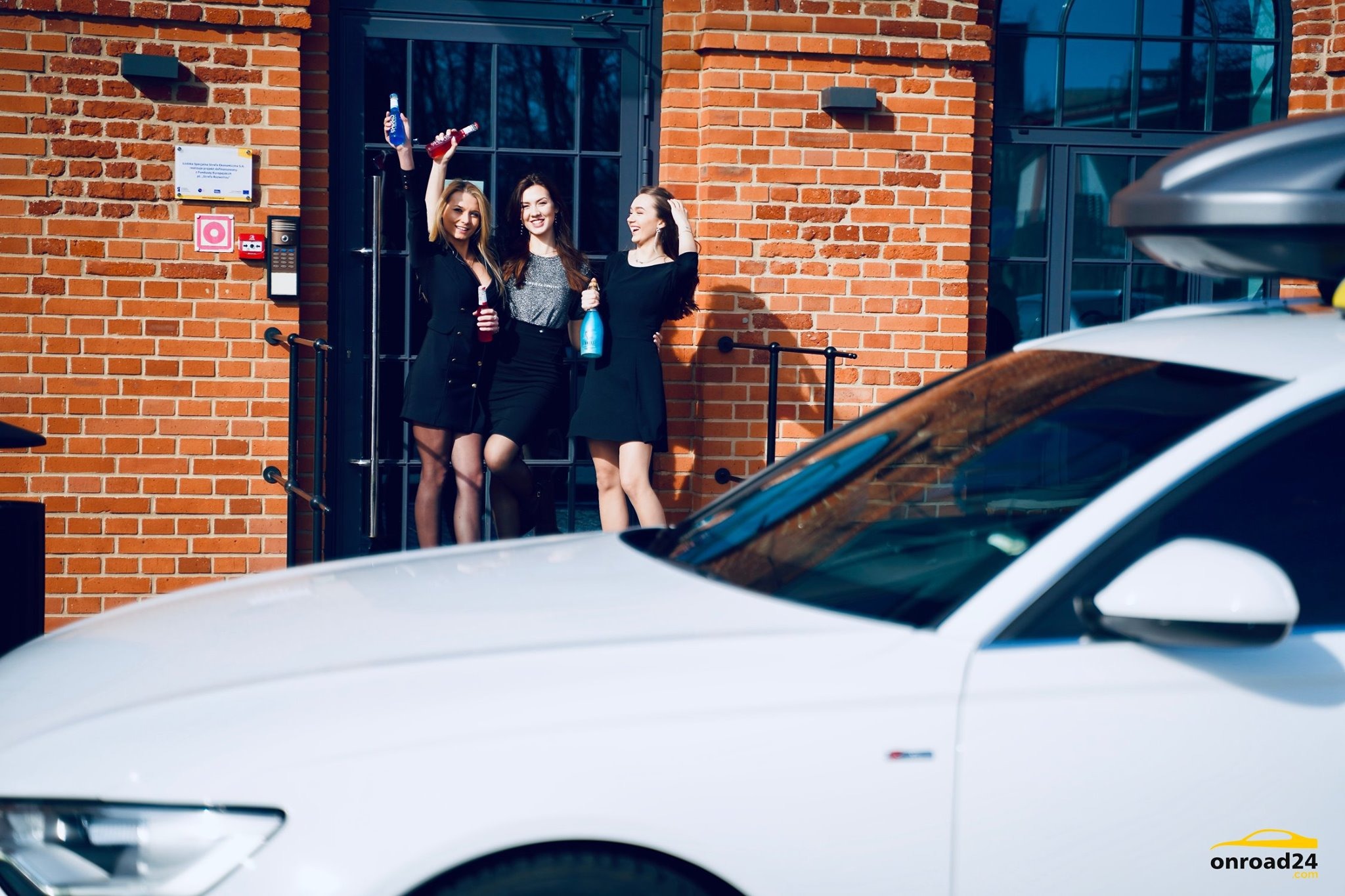 Trzy uśmiechnięte kobiety w czarnych sukienkach świętują z butelkami napojów przed budynkiem z czerwonej cegły, z białym samochodem z bagażnikiem dachowym widocznym na pierwszym planie.