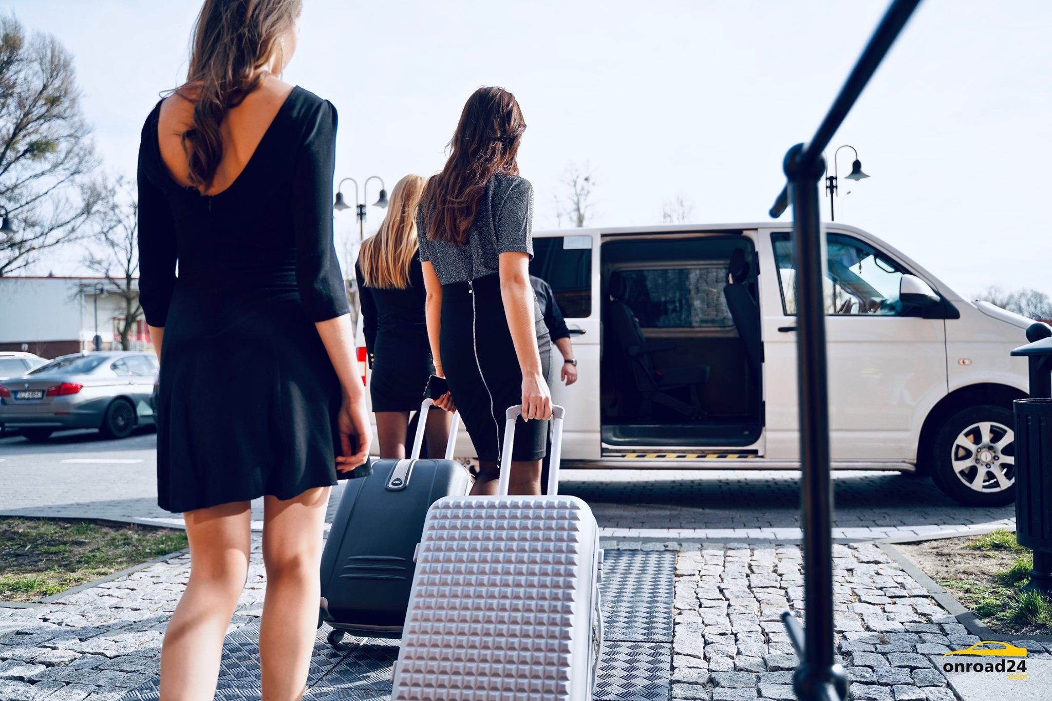 Grupa kobiet z bagażami wsiada do białego busa z otwartymi drzwiami, widok z tyłu, kostka brukowa pod nogami.