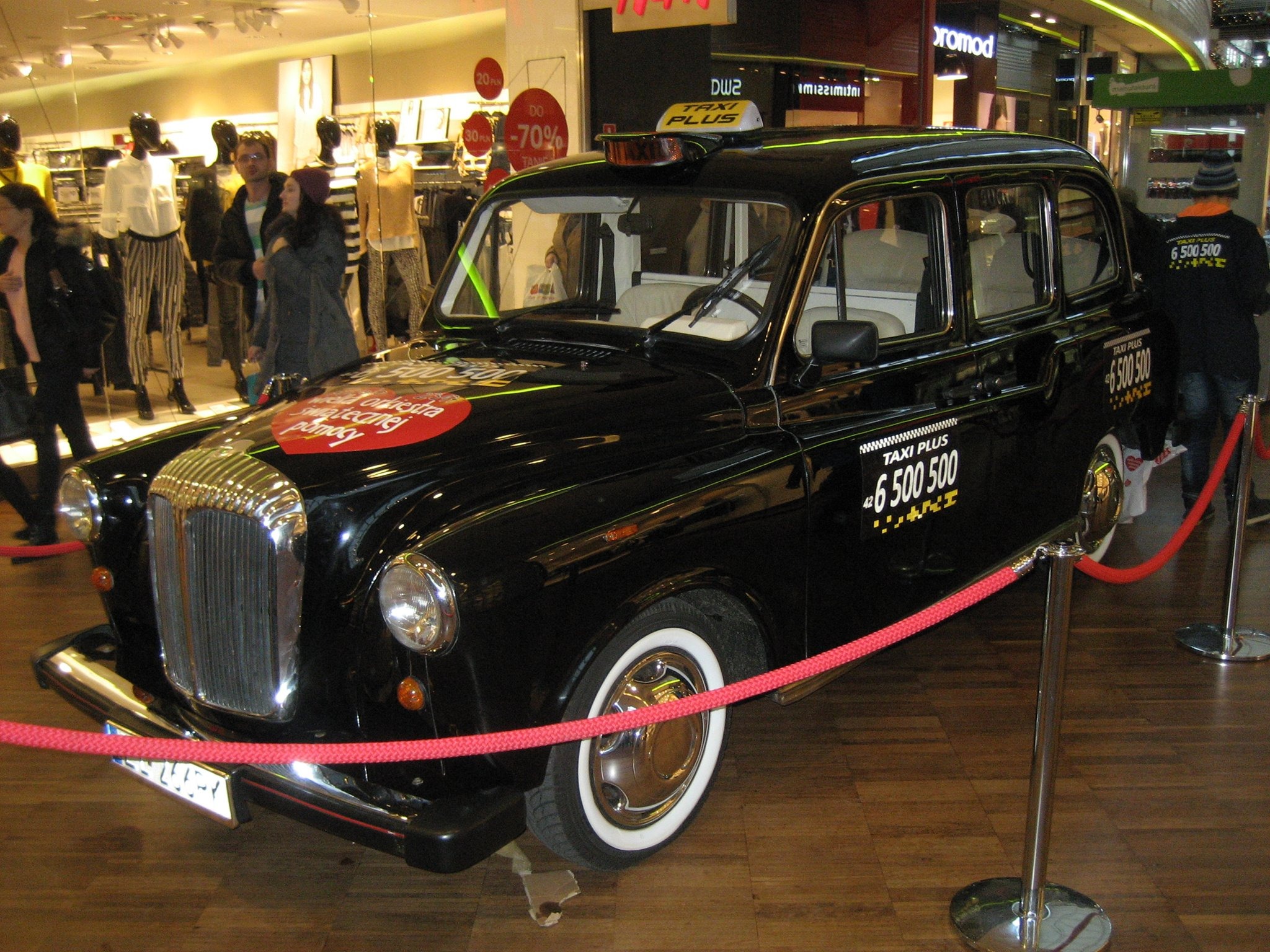Czarna taksówka w stylu retro, z białymi oponami i logo Taxi Plus, wystawiona w centrum handlowym, otoczona czerwonym sznurem odgradzającym.