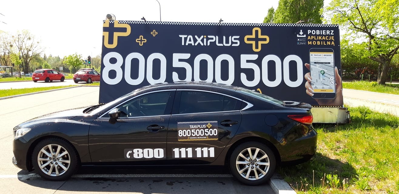 Czarny samochód TaxiPlus z numerami telefonów na karoserii, zaparkowany przed billboardem reklamującym aplikację mobilną.