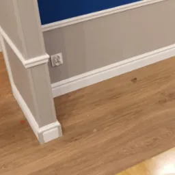 Świeżo pomalowane wnętrze z niebieską ścianą, szarą lamperią i białymi listwami przypodłogowymi, widoczna podłoga z paneli oraz narzędzia malarskie na stole.