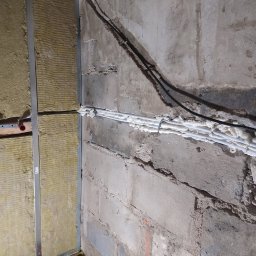 ANDRZEJ RICHTER - Ocieplenie ściany wełną mineralną na ruszcie z profili stalowych, widoczne rury instalacyjne zabezpieczone pianką montażową, surowa ściana z betonu komórkowego.