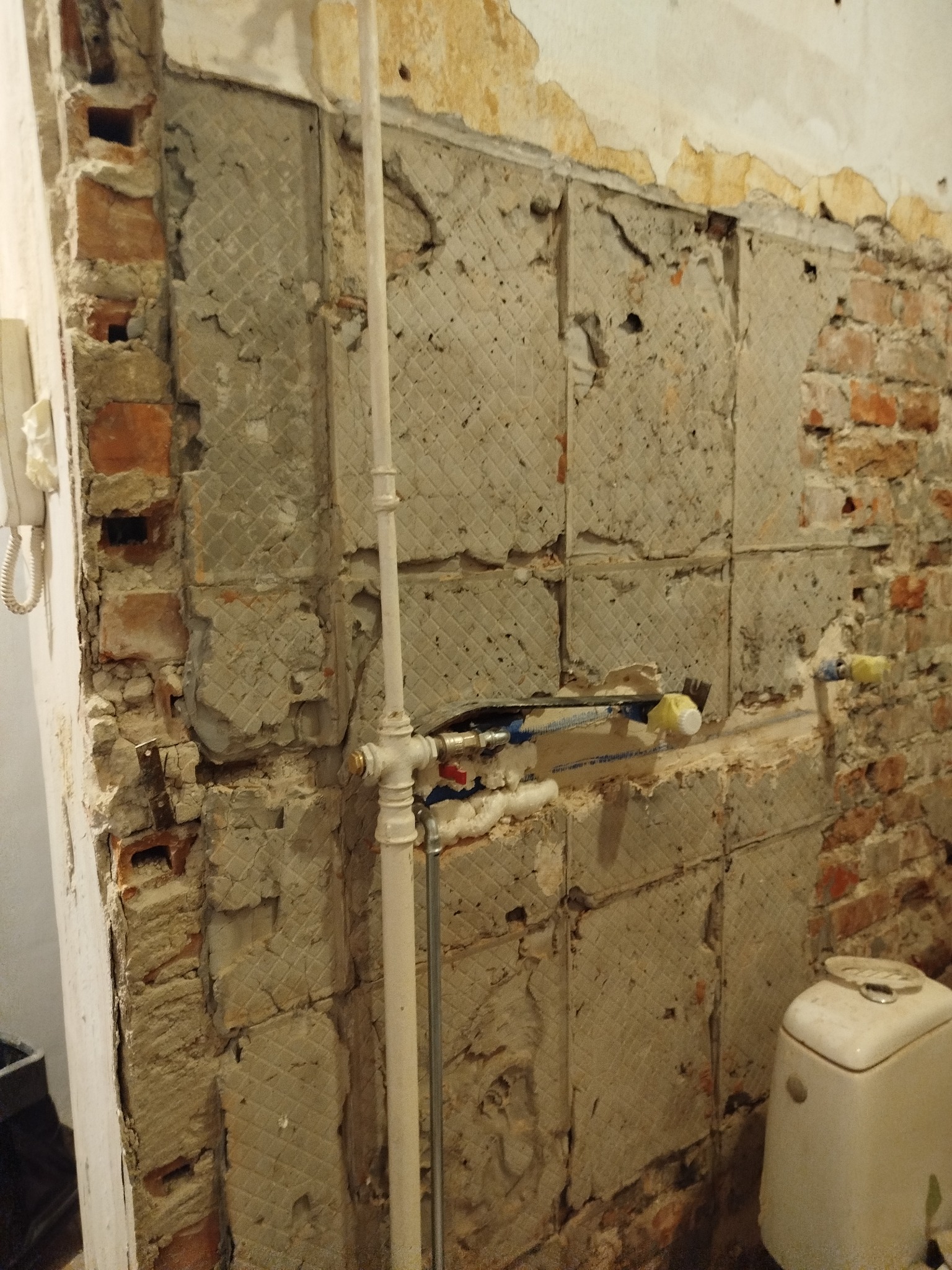 Odsłonięta ściana z fragmentami starej glazury i cegły w trakcie remontu łazienki, widoczne rury instalacji wodnej i fragment toalety.