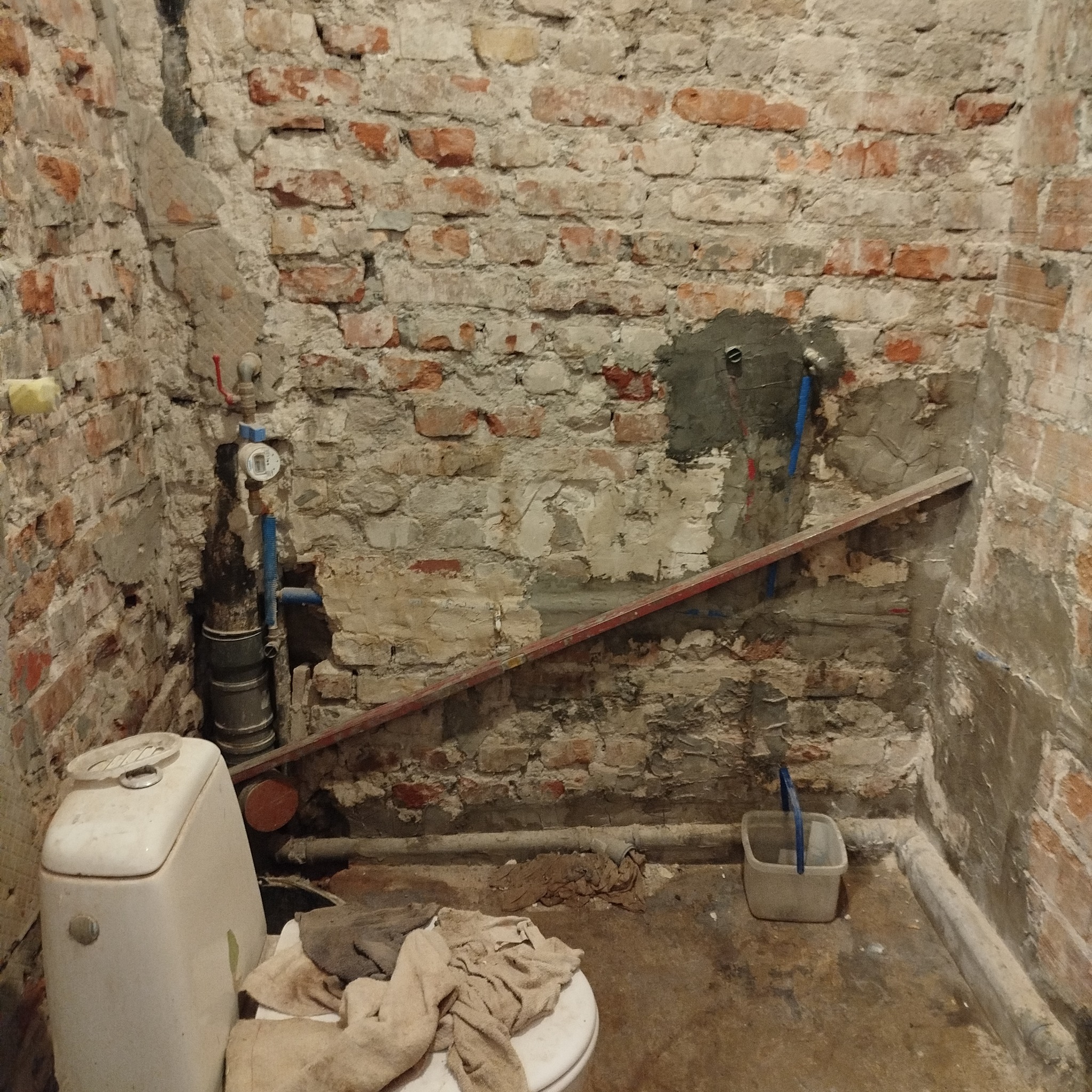 Niedokończona łazienka z odsłoniętą cegłą, instalacją wodną i kanalizacyjną. Widoczne narzędzia i materiały budowlane. Etap prac remontowych w surowym stanie.