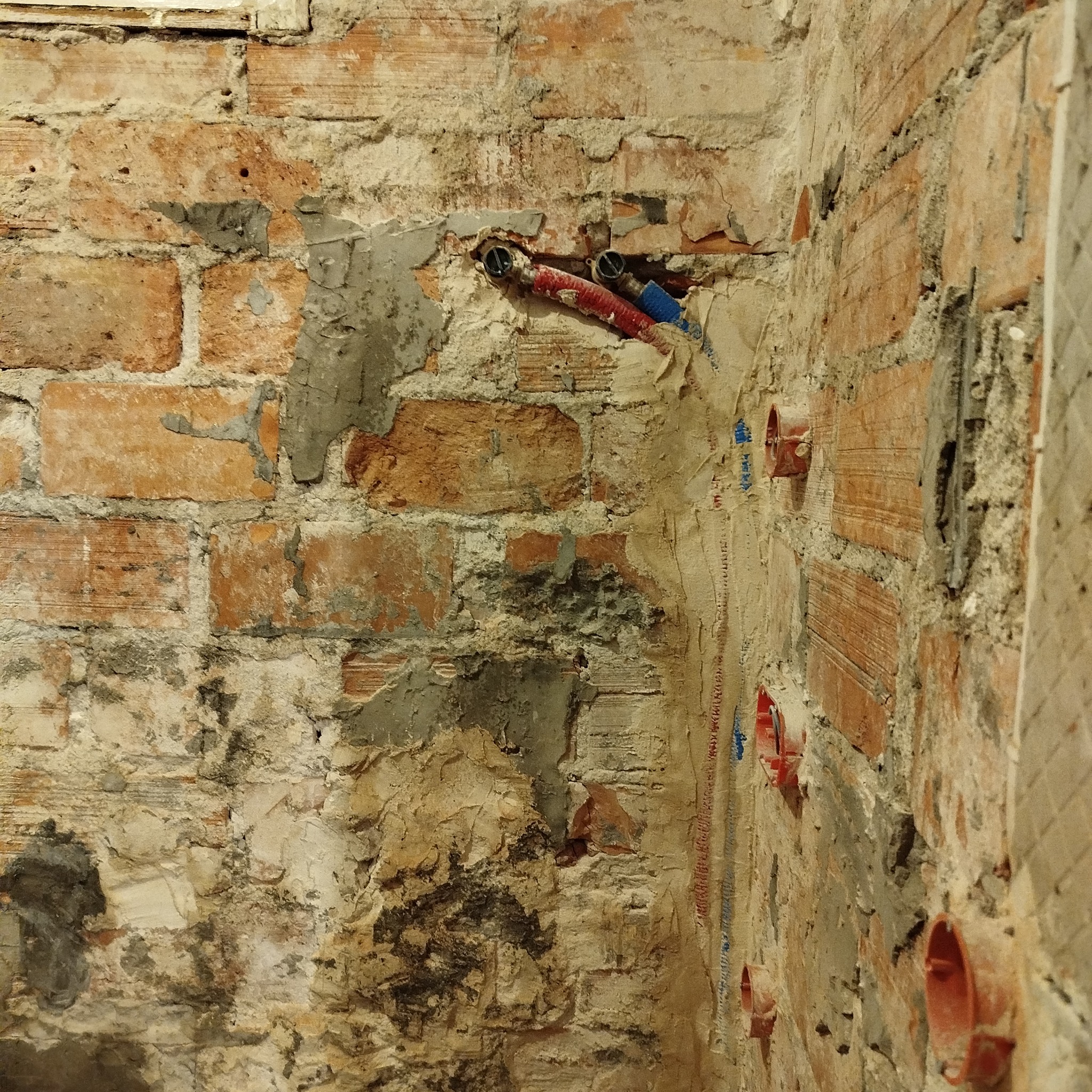 Ceglana ściana w trakcie remontu łazienki z widocznymi rurami instalacji wodnej i elektrycznej, przygotowana do dalszych prac wykończeniowych.