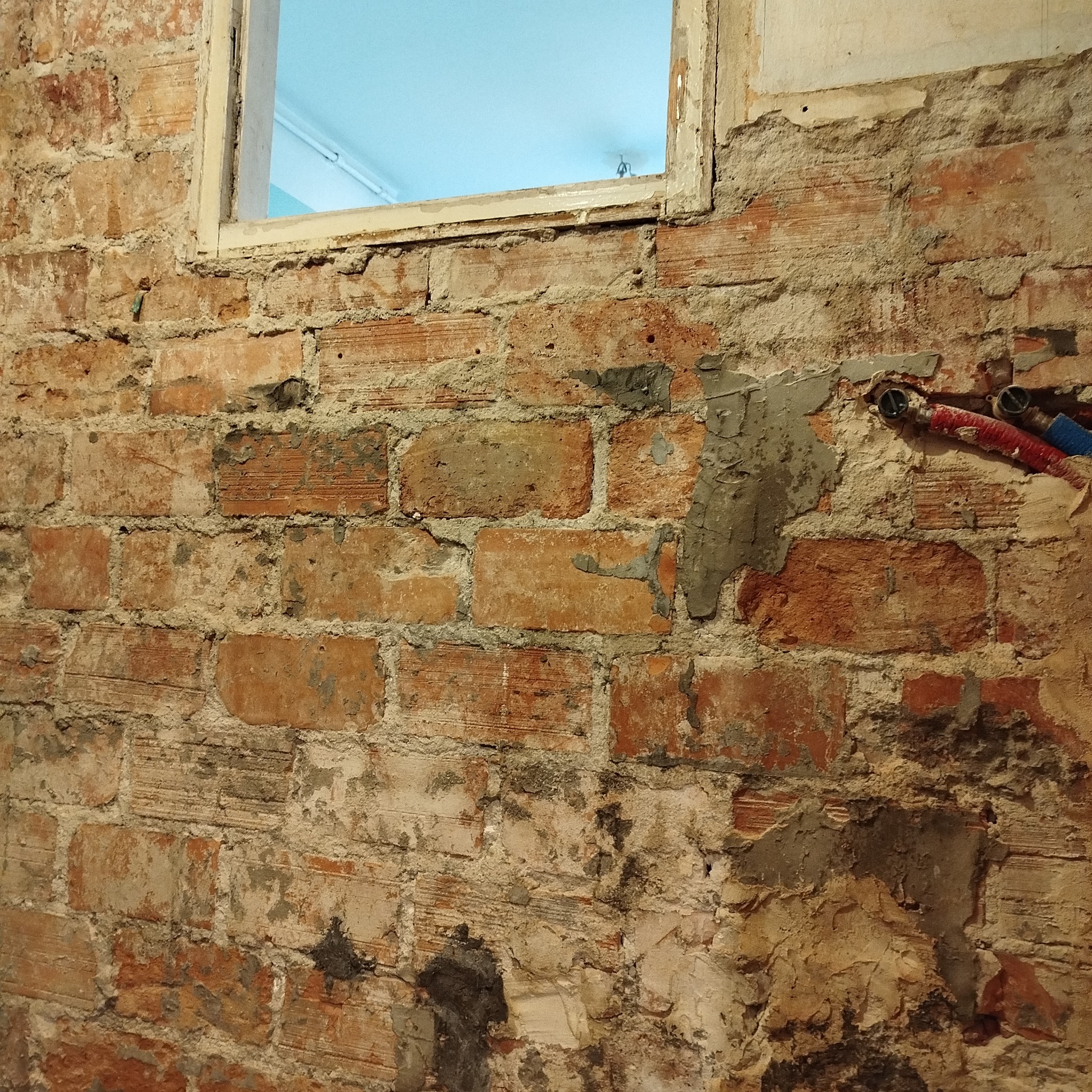 Odsłonięta ceglana ściana z fragmentami starego tynku, widoczne rury instalacji wodnej i okno z widokiem na jasne pomieszczenie. Surowy stan przed pracami wykończeniowymi.