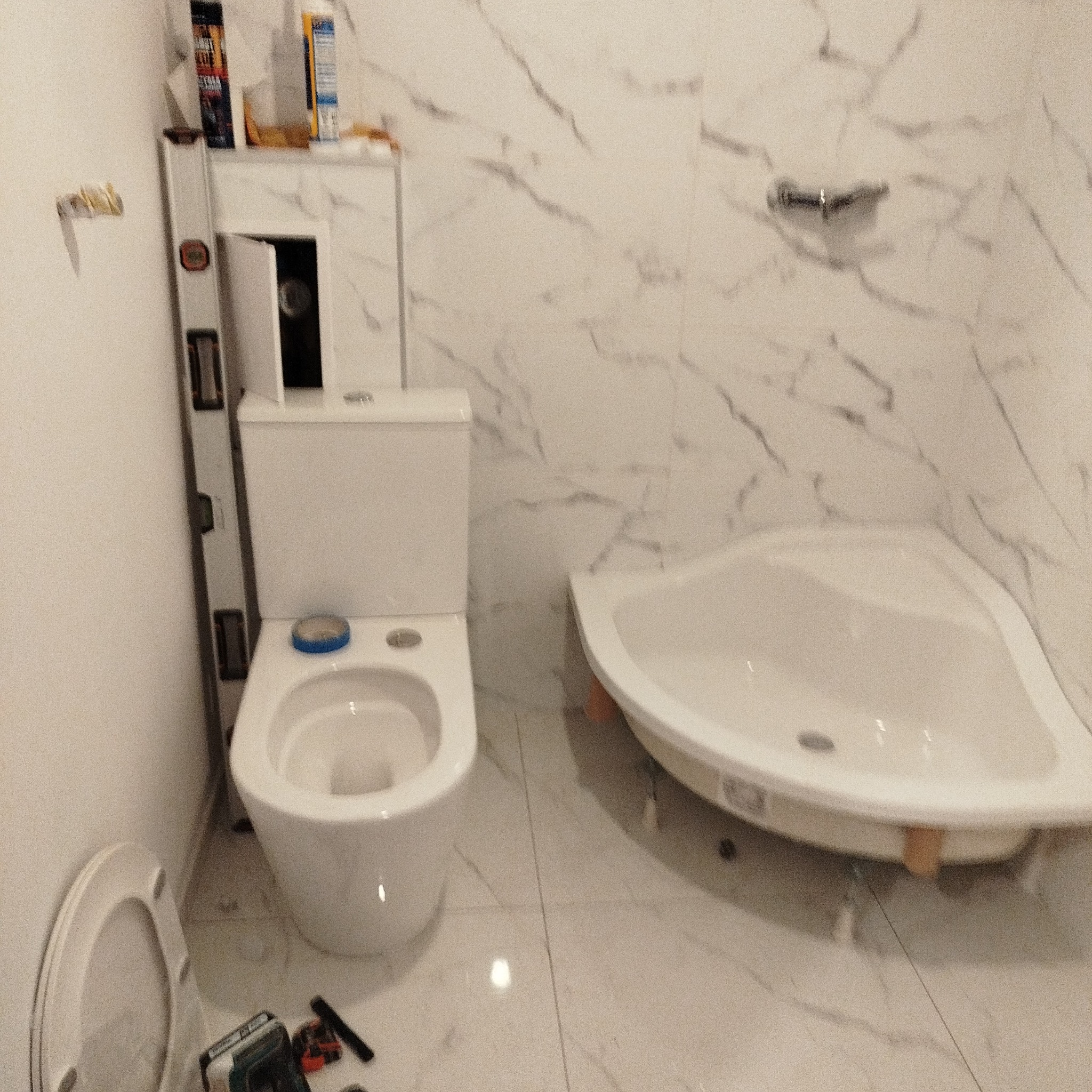 Biała łazienka w trakcie montażu: toaleta z otwartą deską, narożna wanna, poziomica i narzędzia na podłodze. Marmurowe płytki na ścianach.