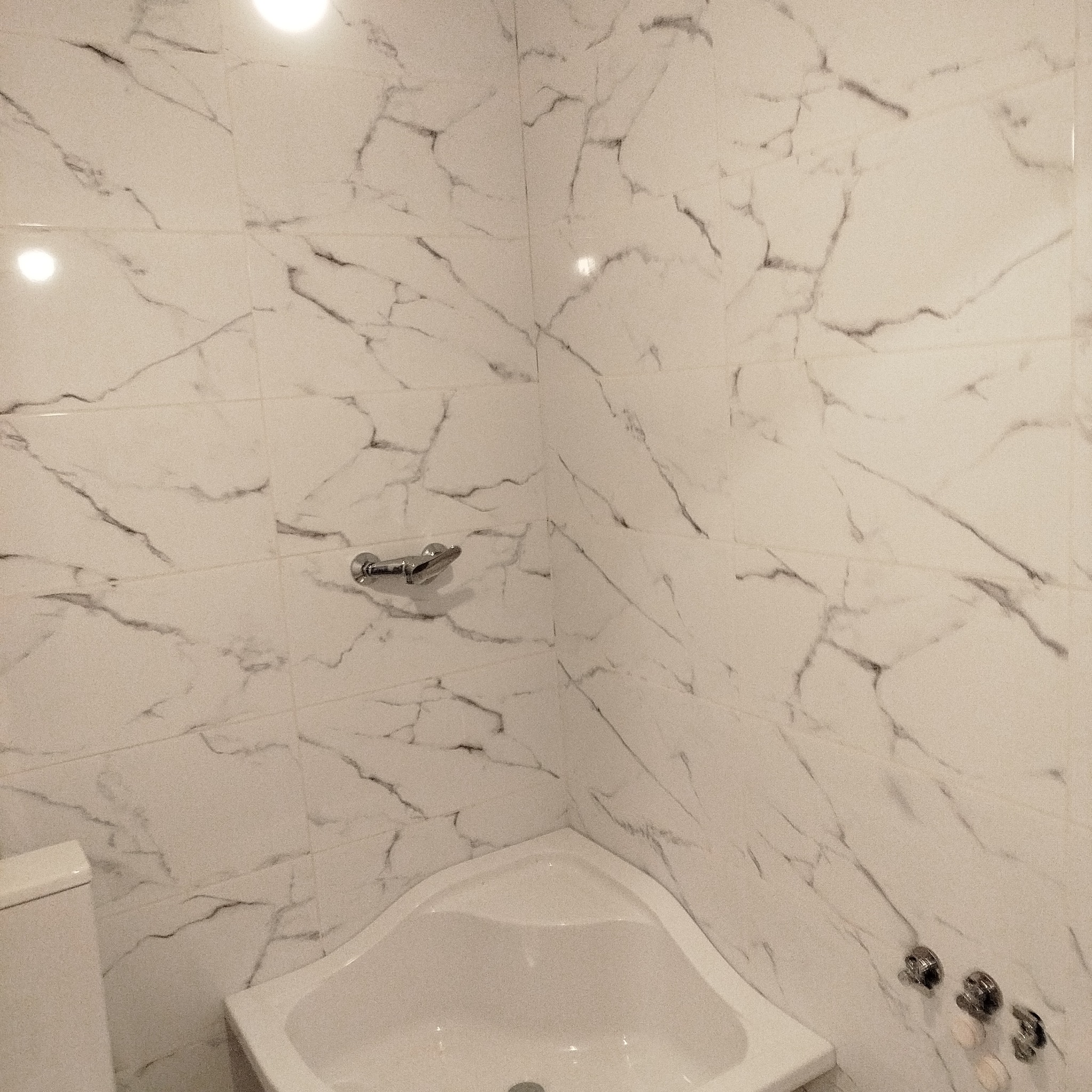 Biała łazienka z marmurowymi płytkami, narożną wanną i chromowaną armaturą. Widoczne detale wykończenia, takie jak uchwyt na ręcznik i zawory.