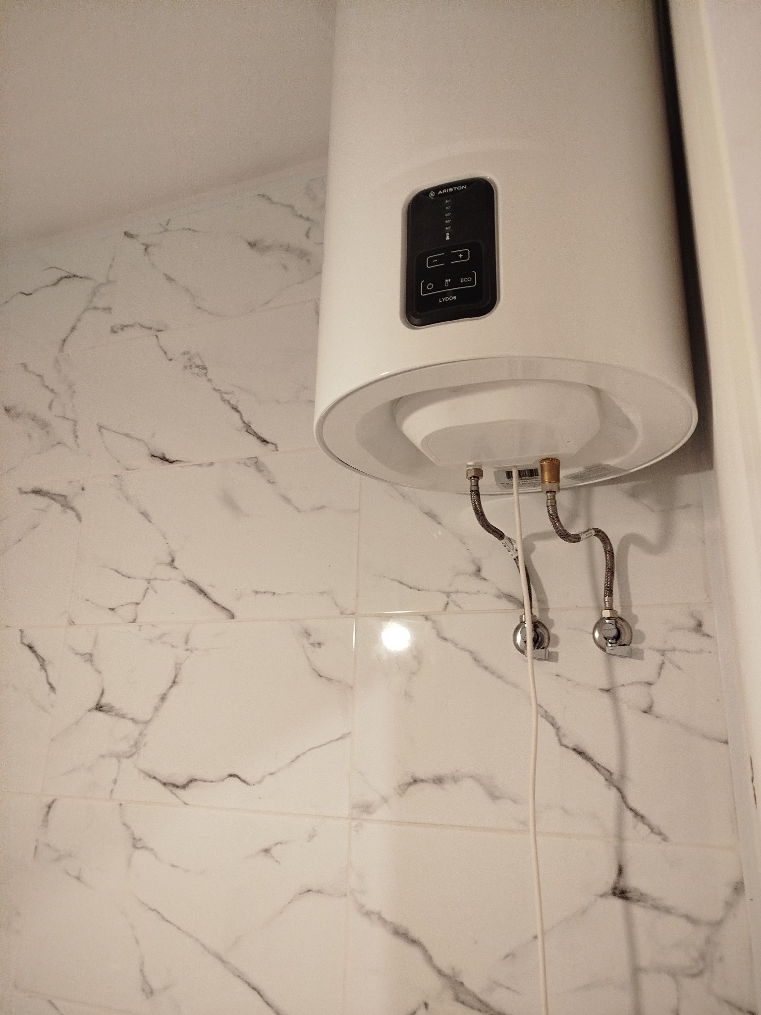 Biały, cylindryczny bojler Ariston zamontowany na ścianie z marmurowych płytek w łazience. Widoczne przyłącza wodne i panel sterowania bojlera.