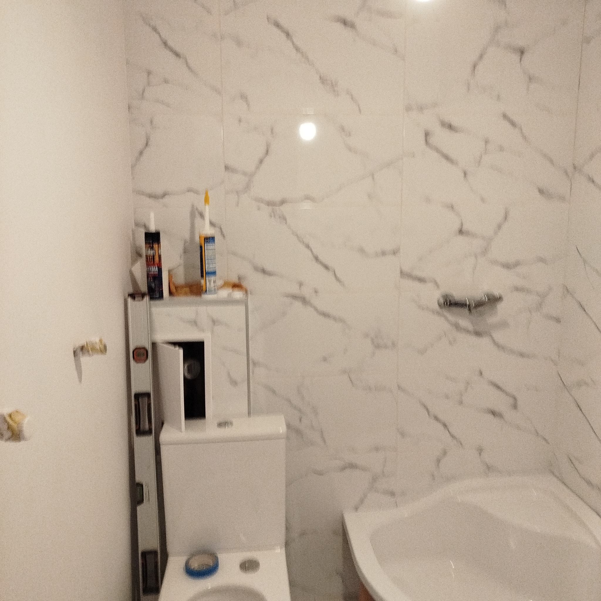 Remont łazienki: białe płytki imitujące marmur na ścianach, częściowo zamontowana toaleta, poziomica i silikony. Widoczny fragment wanny.