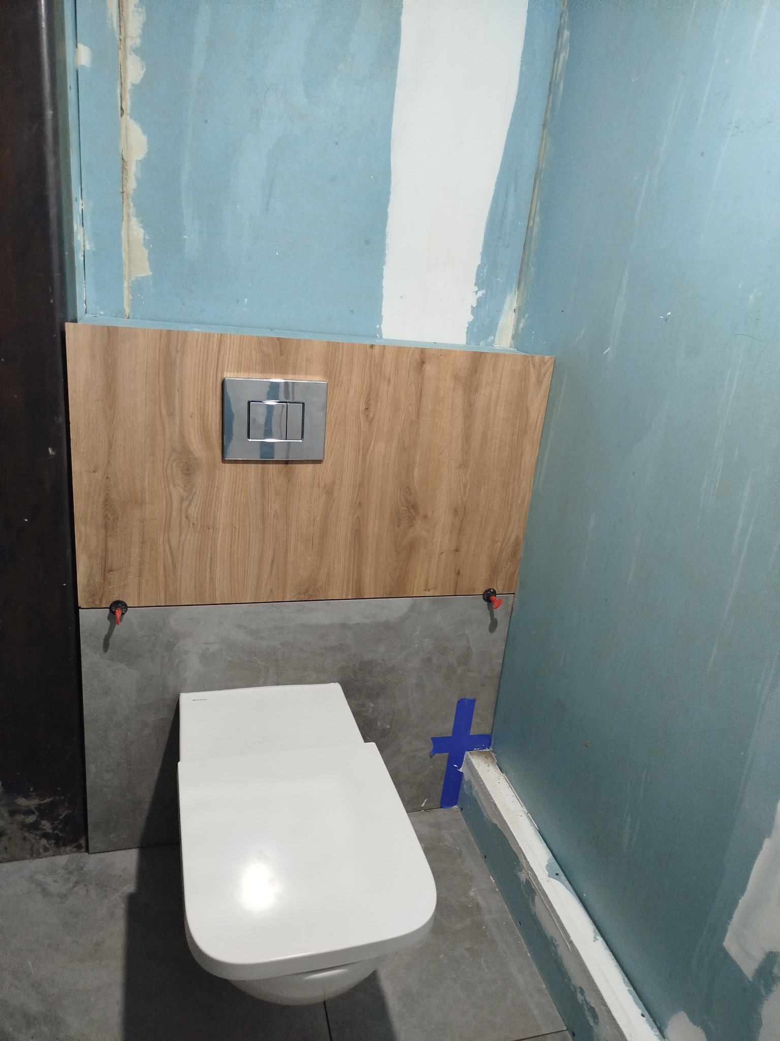 Montaż stelaża podtynkowego WC z drewnopodobną obudową i przyciskiem spłukującym na tle surowej ściany w trakcie prac wykończeniowych w łazience.