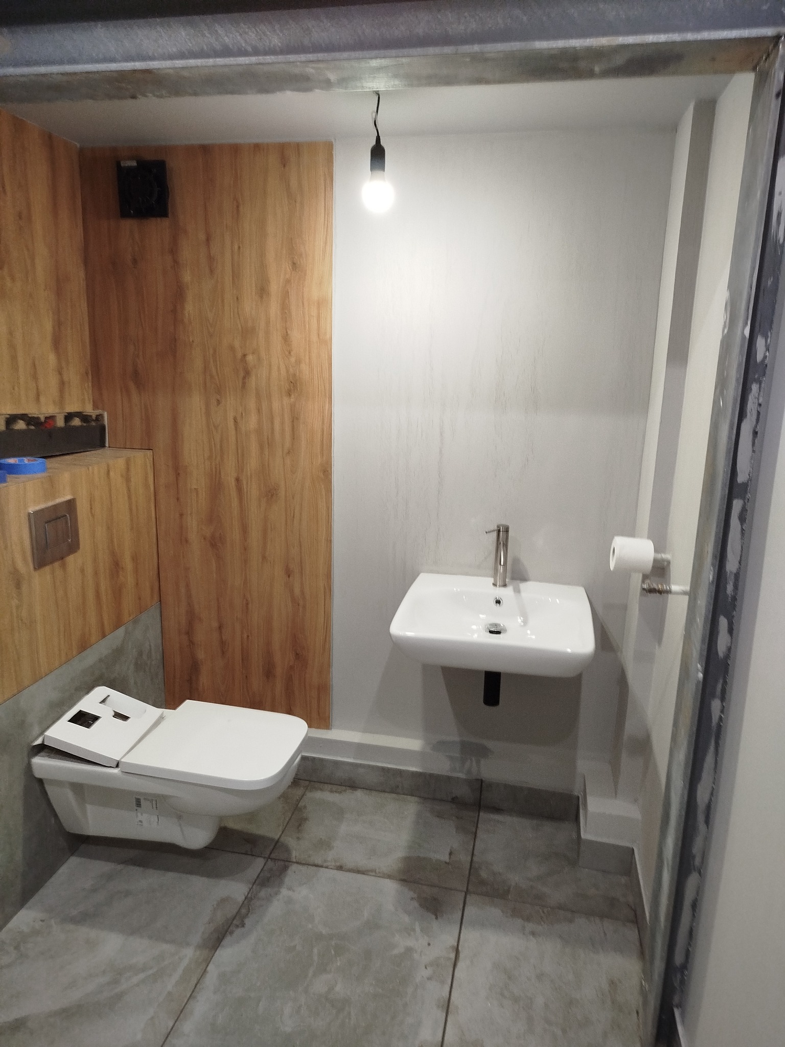 Nowoczesna łazienka po remoncie: biała umywalka i toaleta z deską elektroniczną na tle drewnopodobnych paneli ściennych i szarej podłogi z płytek.